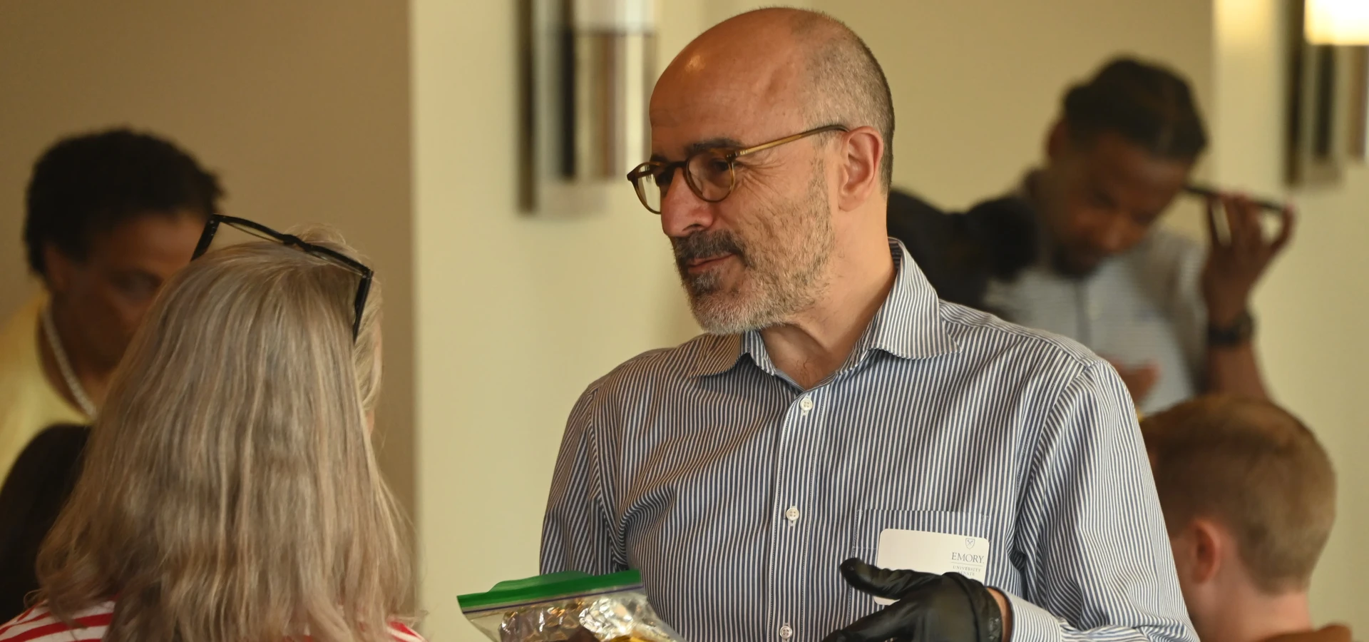 A Senate member in food preparation gloves holds a wrapped sandwich while conversing with a colleague